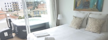 A hotel room with a bed, pillows, a window with curtains, a painting on the wall, and a view of buildings outside.