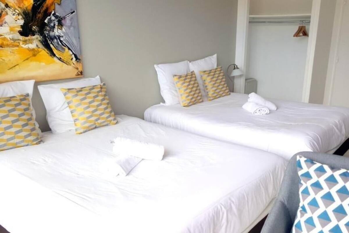 A hotel room with two beds, each with white linens and patterned pillows. There is a painting on the wall, a lamp on the nightstand, and a purple chair.