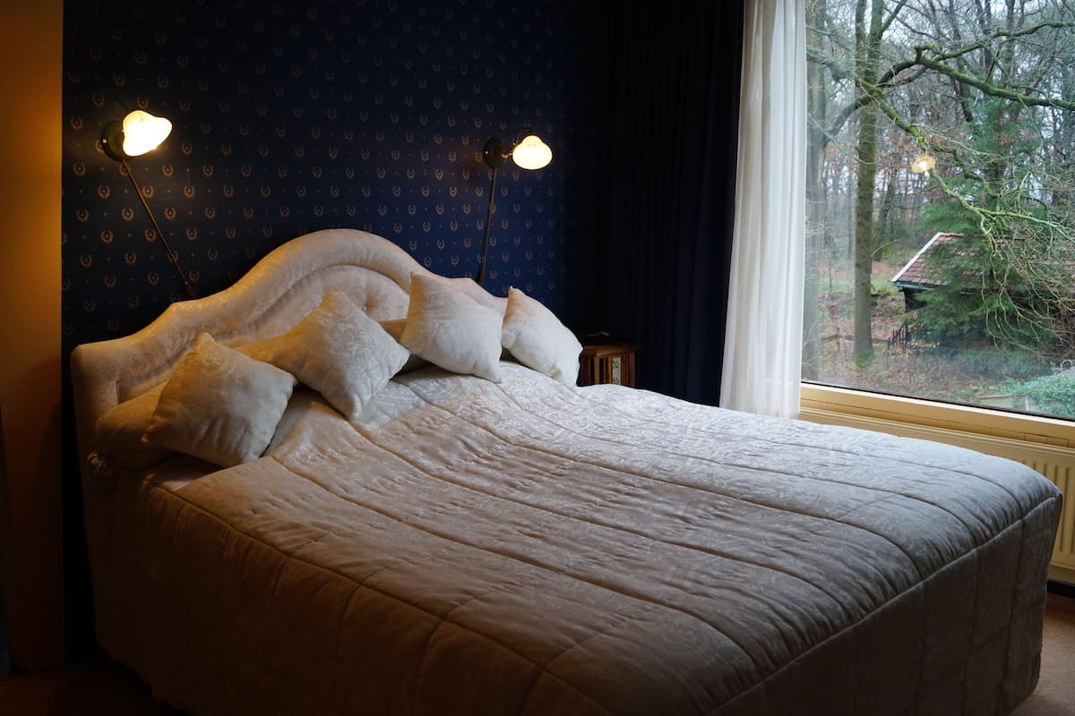 Una cama con colcha y almohadas blancas, pared azul oscuro con diseño y ventana con vista a árboles y una casa.