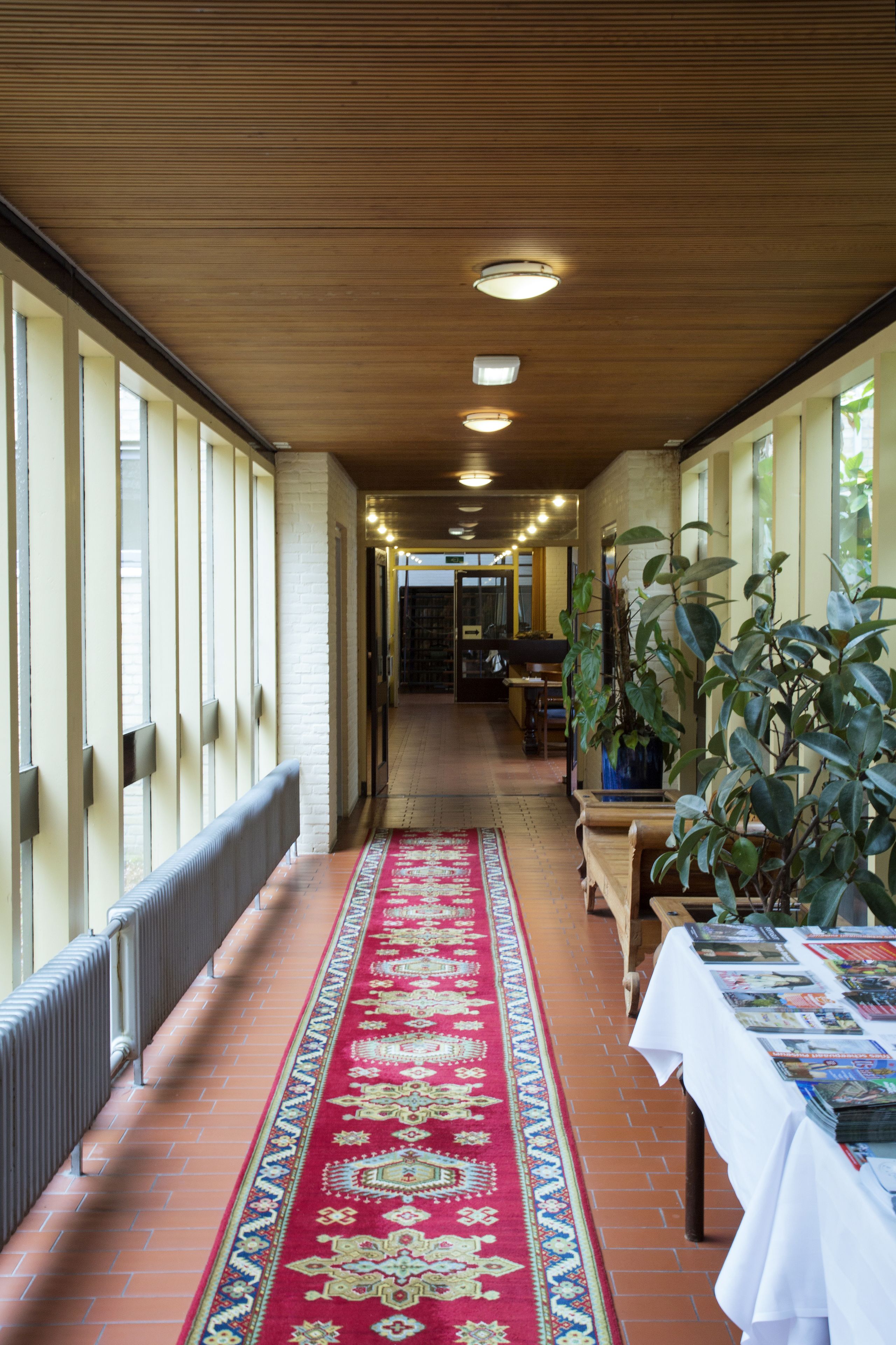A corridor with a red patterned carpet, wooden ceiling, and large windows on one side.