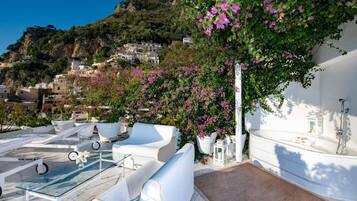 A rooftop terrace with white outdoor furniture, a glass table, and a white bathtub.