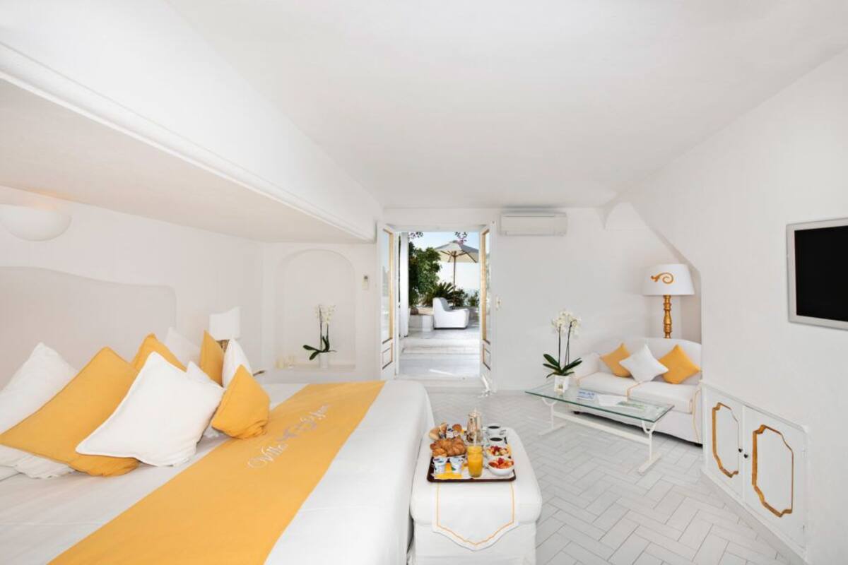 A bright, white hotel room with a bed, a breakfast tray, a sofa, and a view of the outdoor area.