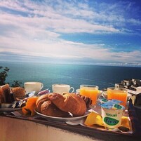 Bandeja de desayuno con croissants, café, jugo y yogur en un balcón con vista al mar.