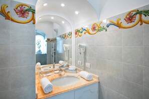 A bathroom with a marble sink, a mirror, a hairdryer, and decorative tiles. - Villa Yiara (Positano)