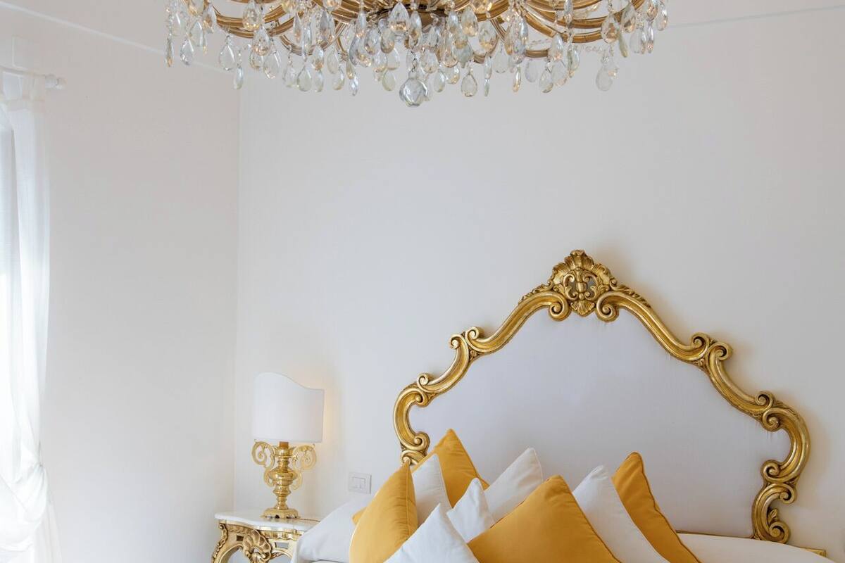 A luxurious bedroom with a large ornate chandelier, a gold-framed headboard, and a bed with white and yellow bedding.