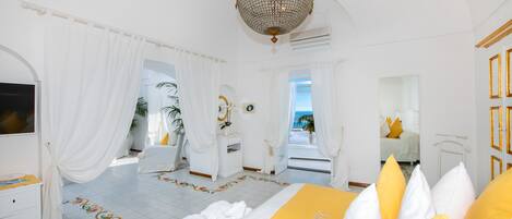 A bright, white hotel room with a large bed, a chandelier, and a view of the sea.