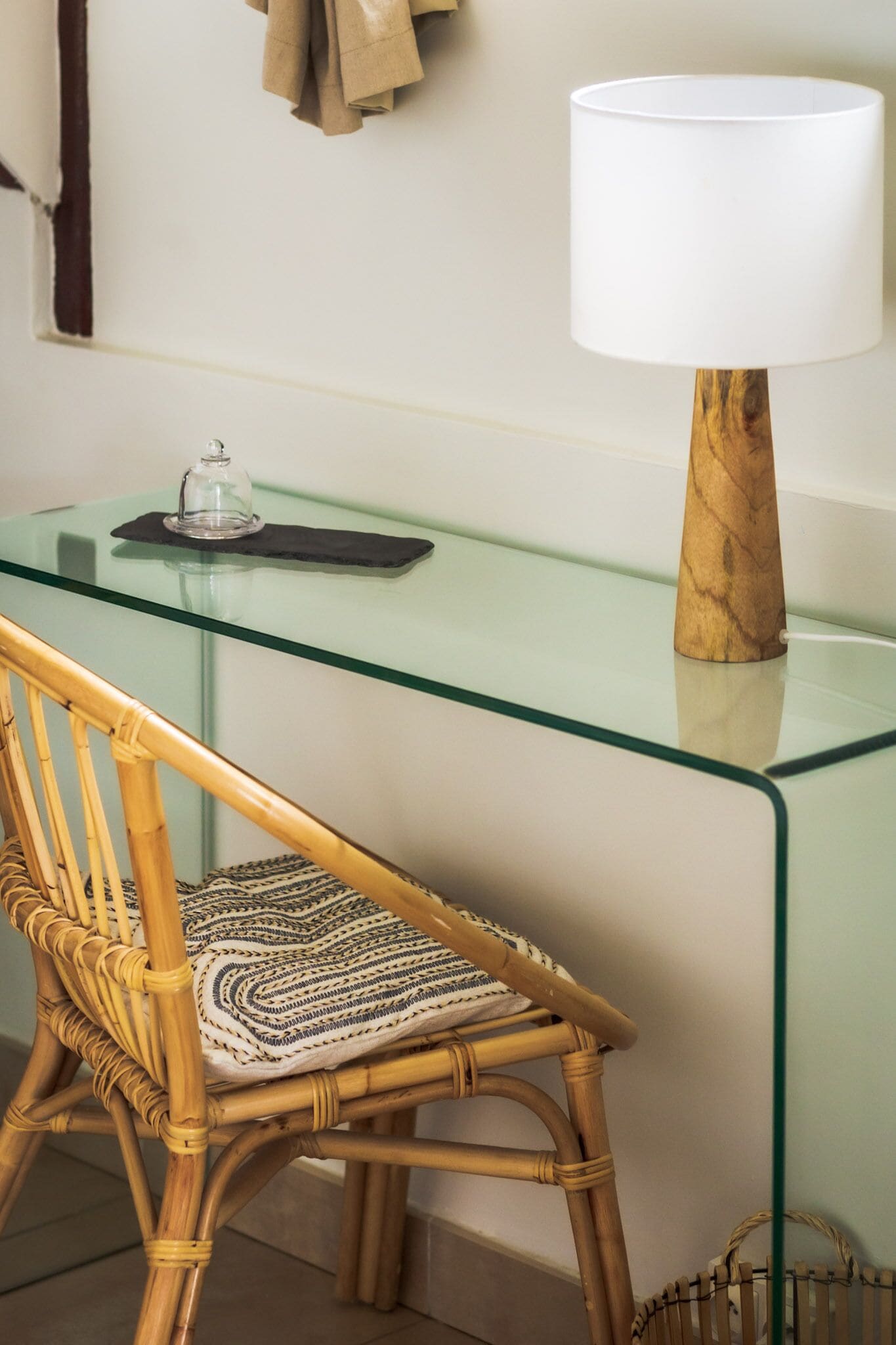 Un bureau à plateau en verre, une lampe en bois, une chaise en osier et un petit récipient en verre posé sur le bureau.