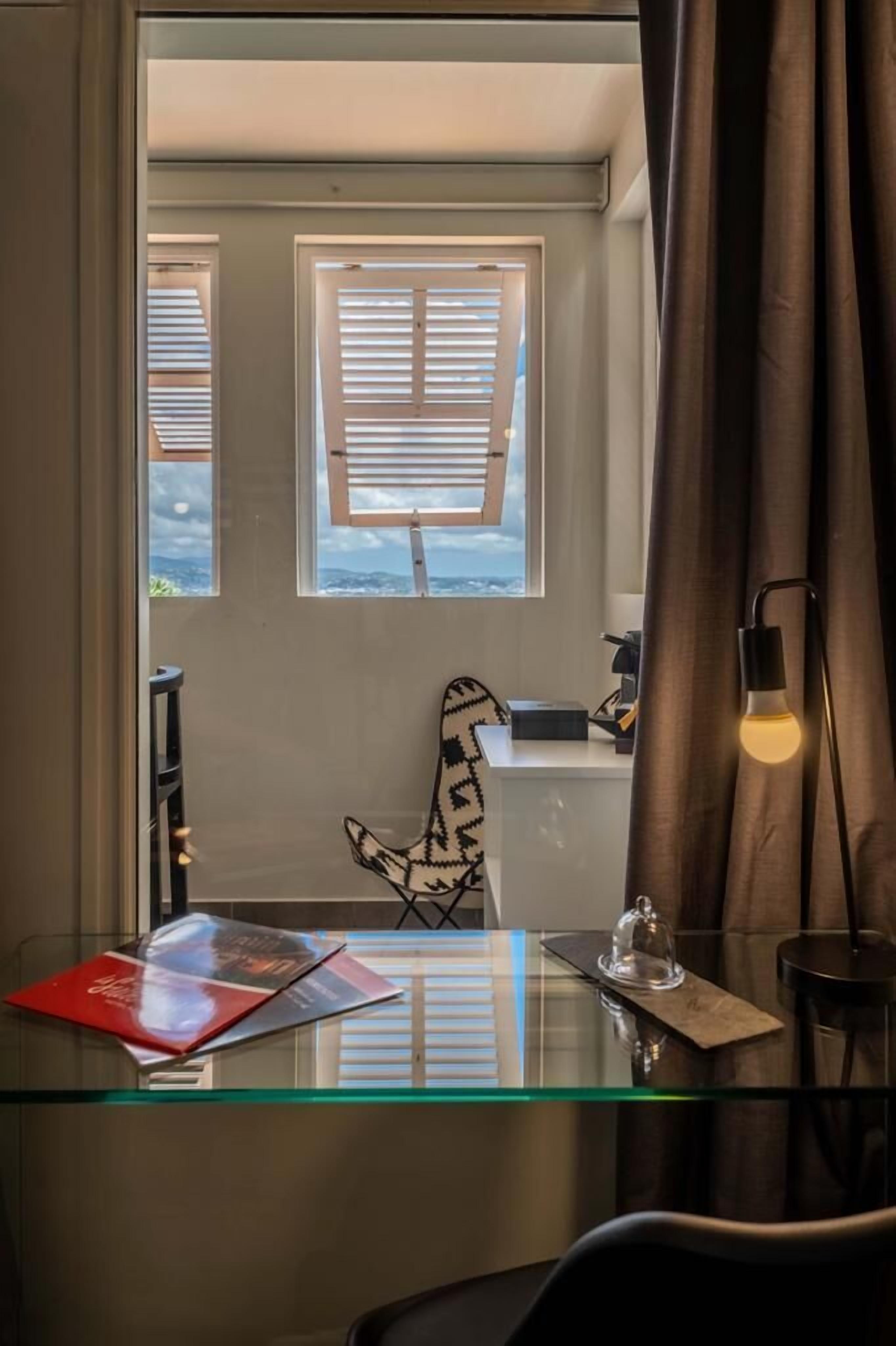 Una habitación con un escritorio de vidrio, una silla, una mesita con una campana y una ventana con persianas.