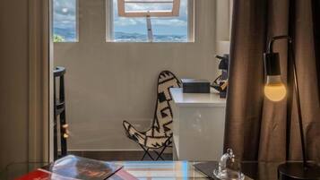 Una habitación con un escritorio de vidrio, una silla, una mesita con una campana y una ventana con persianas.