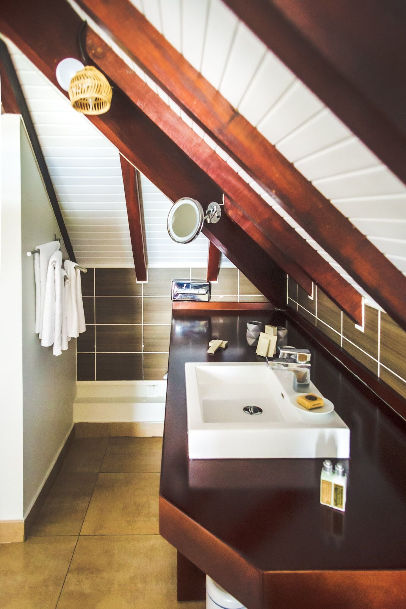 A bathroom with a wooden vanity, a white sink, and a mirror. There are towels hanging on the wall and a small shelf with toiletries.
