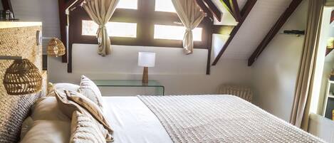Una camera da letto con soffitto spiovente, un letto grande con copripiumino e cuscini bianchi, un comodino con una lampada e una finestra con tende.