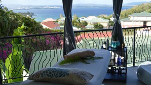 A balcony with a view of the sea and houses, a bed with a pillow and a plant, and a small table with bottles and a vase. - La Suite Villa (Trois-Ilets)