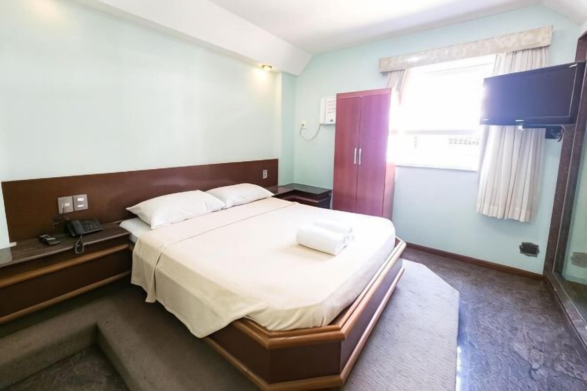 A hotel room with a bed, bedside table, lamp, and a window with curtains.