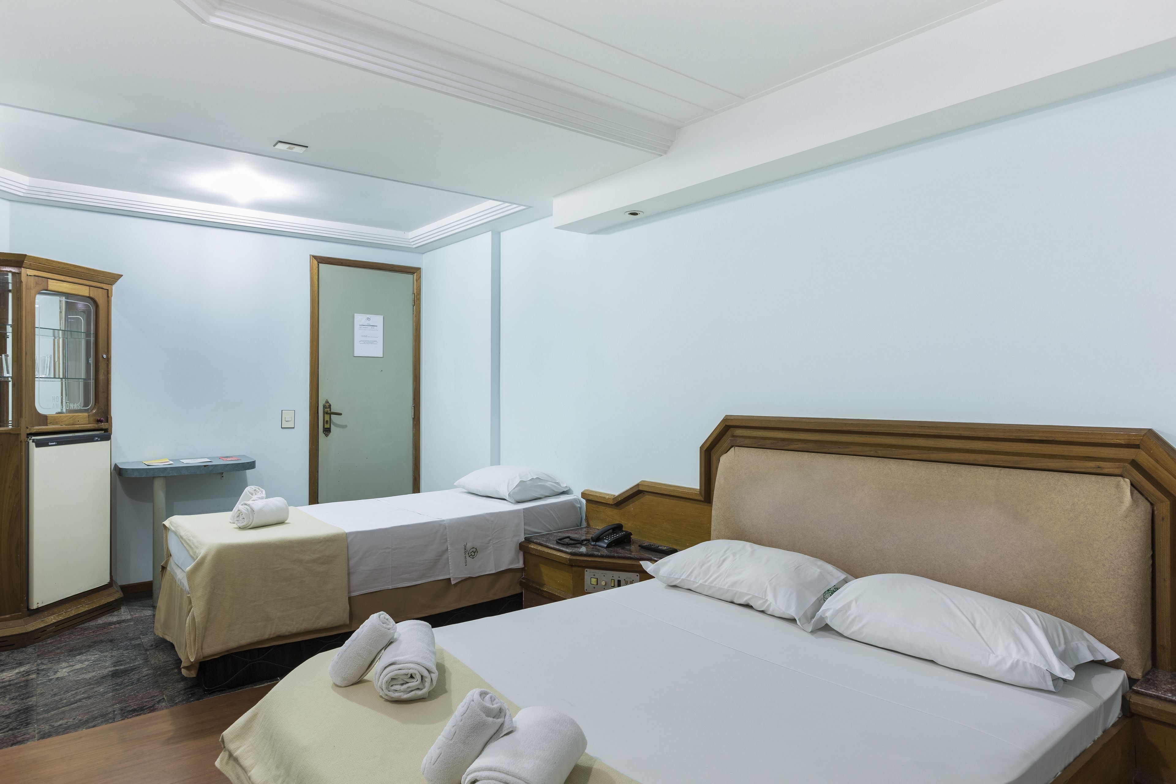 Quarto de hotel com duas camas de solteiro, cabeceira de madeira, pequena escrivaninha com cadeira, espelho e porta com placa.