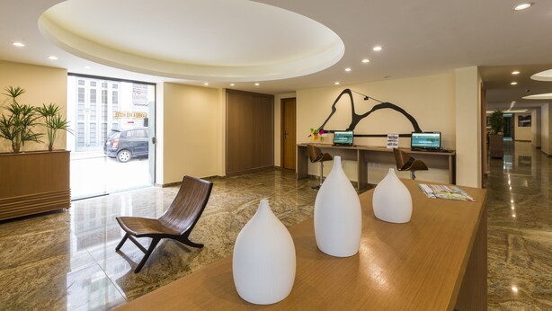 Un área de recepción con un mostrador de madera, tres jarrones blancos, una silla marrón y un escritorio con una computadora y una planta.