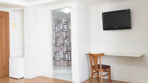 Una habitación con puerta de madera, un televisor blanco montado en la pared, una silla de madera y una cortina de ducha estampada.