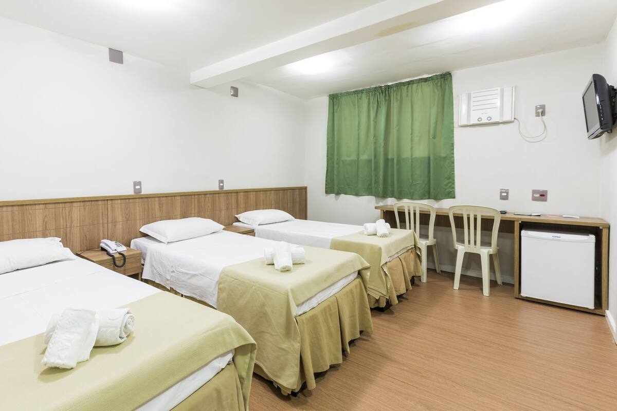Habitació d'hotel amb quatre llits individuals, una taula de nit amb cadira, un refrigerador petit i una televisió penjada a la paret.