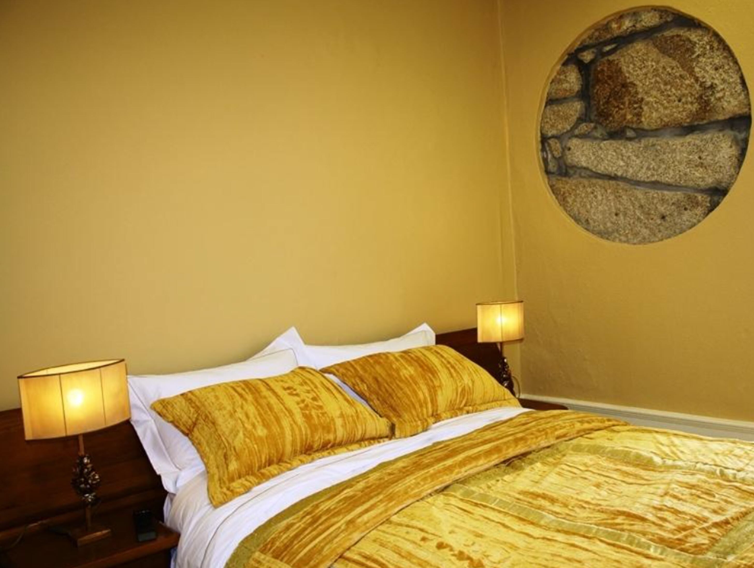A bed with a gold-colored bedspread and pillows, two bedside lamps, and a stone wall feature.