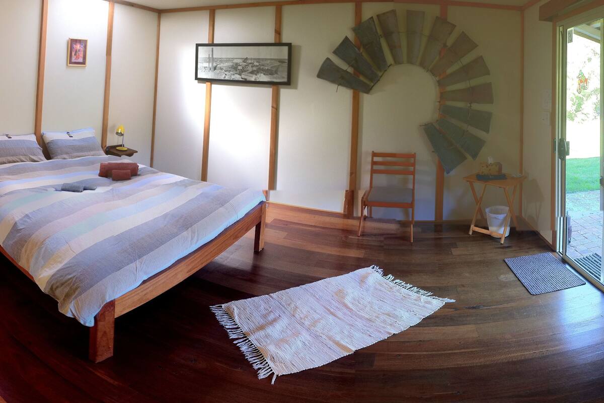 A bedroom with a bed, wooden flooring, a wall-mounted fan, and a small table with a chair.