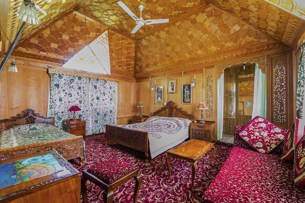 Quarto tradicional com teto de madeira, cama com colcha estampada, mesa de madeira e chão coberto por um tapete vermelho.