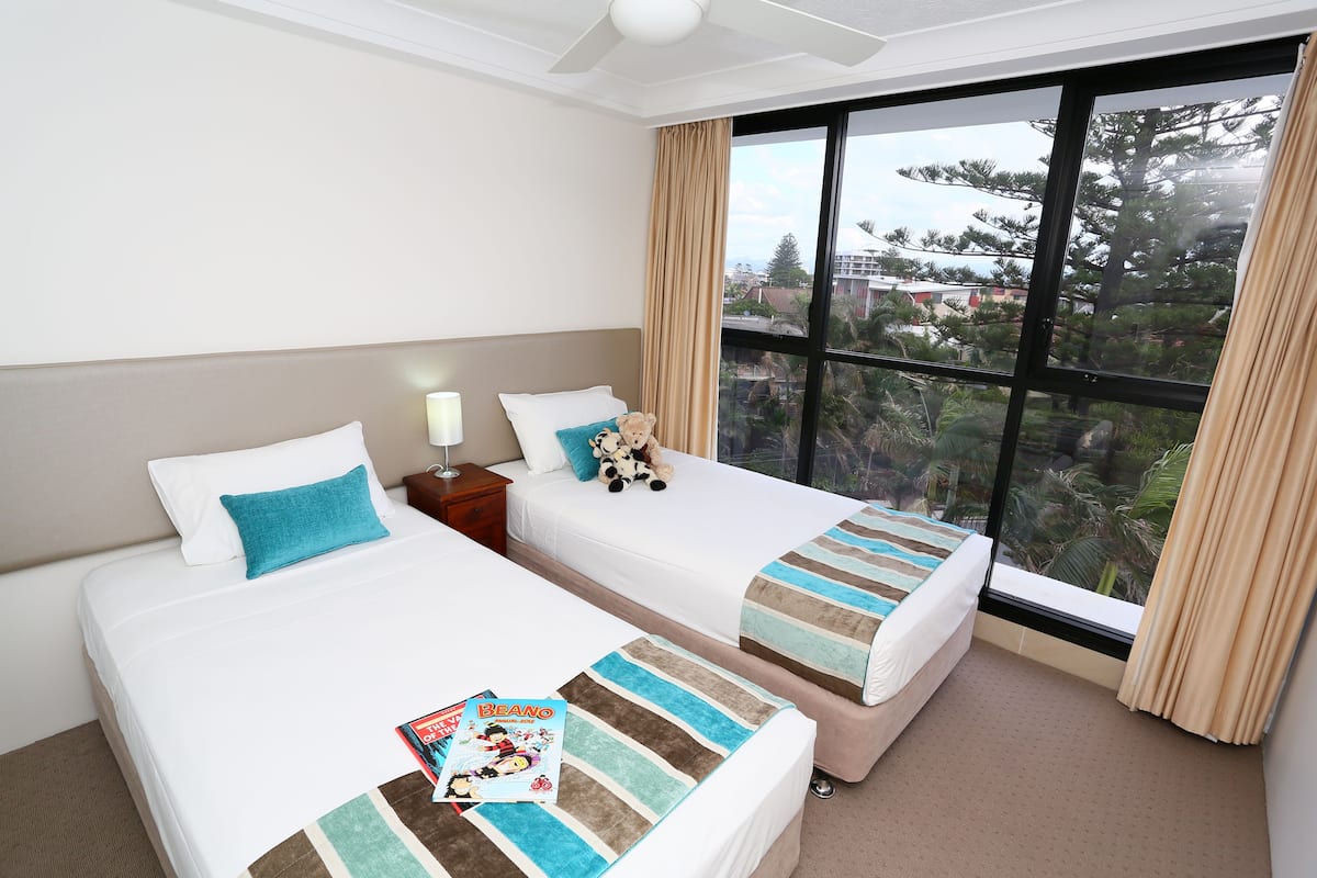 A hotel room with two beds, a large window with curtains, and a view of trees and buildings outside.