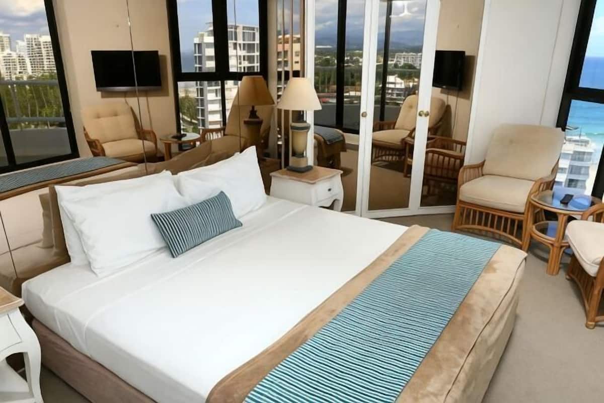 A hotel room with a large bed, a TV, a balcony with chairs, and a view of the city.