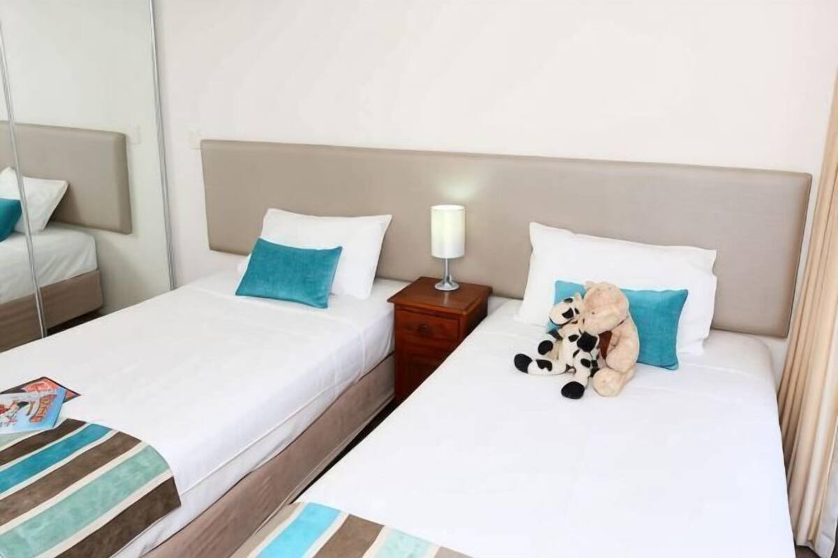 A hotel room with two single beds, a nightstand with a lamp, and a teddy bear on one bed.