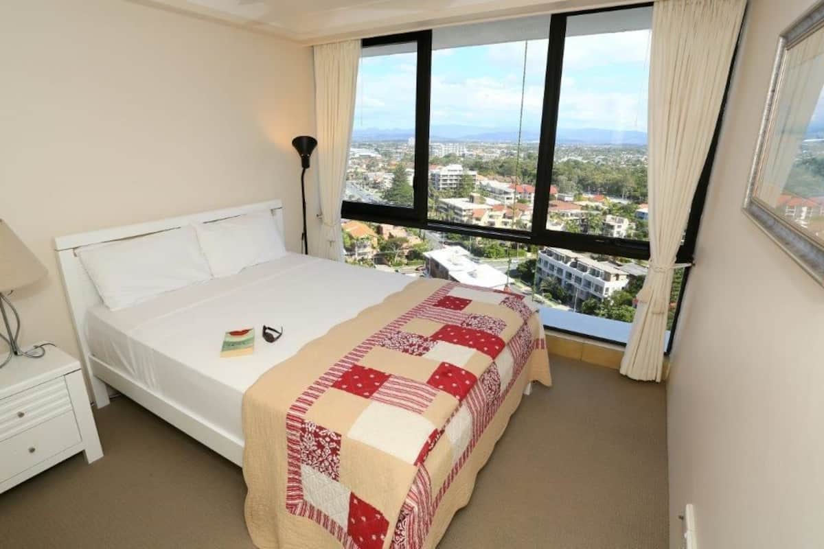 A hotel room with a bed, a nightstand, a lamp, and a large window offering a city view.