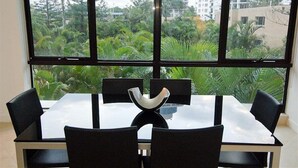 A dining area with a glass table and black chairs, large windows with a view of greenery and buildings.