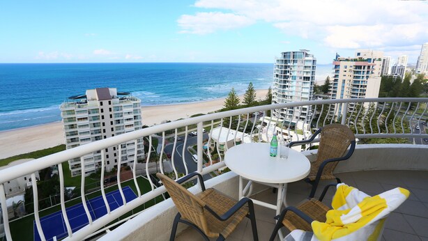 A balcony with wicker chairs and a table, overlooking a beach and ocean with high-rise buildings in the background.