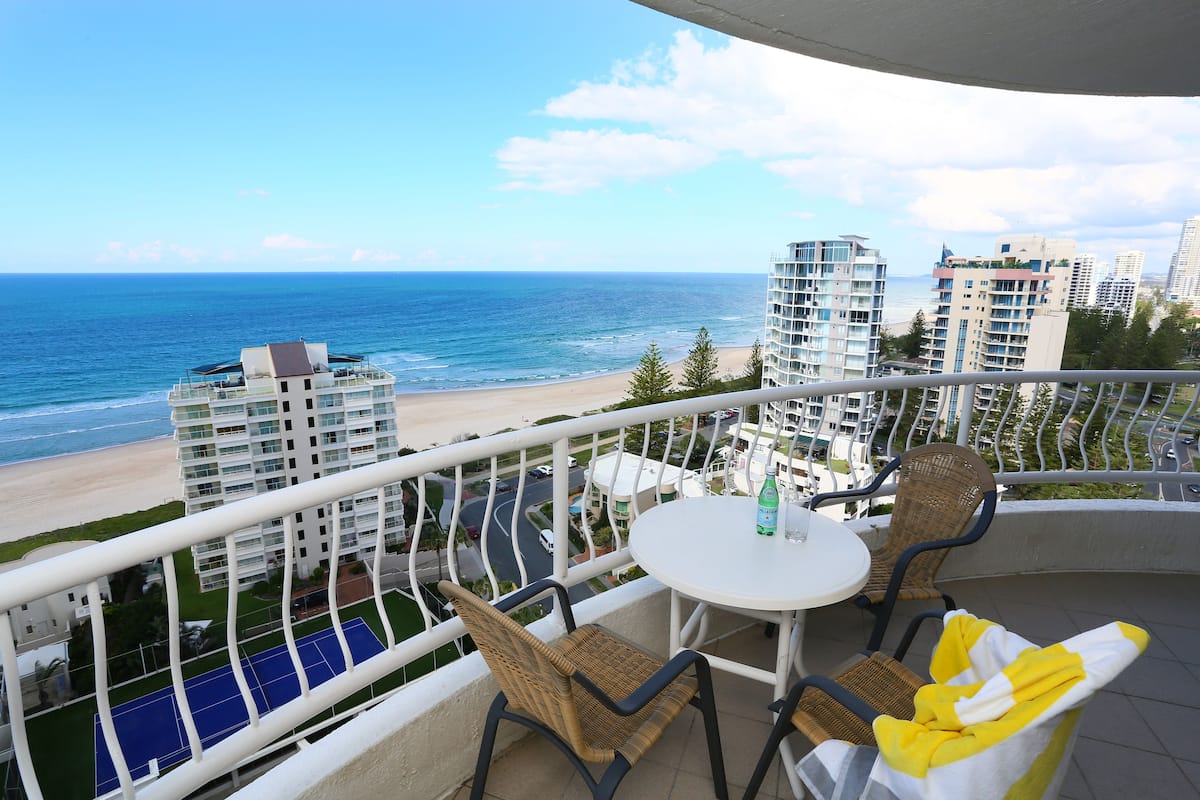A balcony with wicker chairs and a table, overlooking a beach and ocean with high-rise buildings in the background.