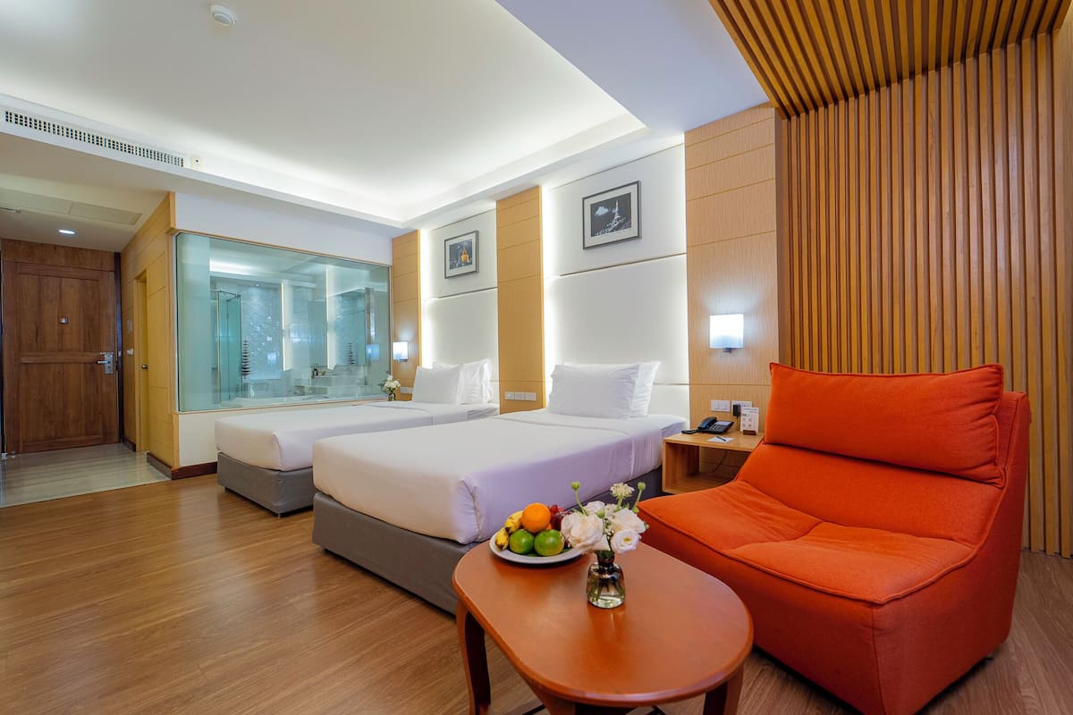 Hotelkamer met twee bedden, een houten vloer, een douchecabine van glas en een klein tafeltje met een schaal fruit en bloemen.