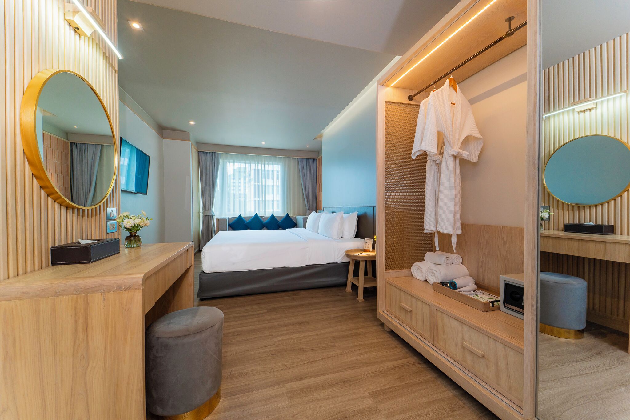 Una habitación de hotel moderna con un escritorio de madera, una cama grande con almohadillas azules, un tocador con espejo y un vestidor con una bata colgada.