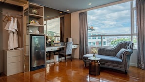 Una habitación de hotel moderna con piso de madera, un sofá gris, un escritorio con una silla, un armario y un ventanal con vistas a la ciudad.