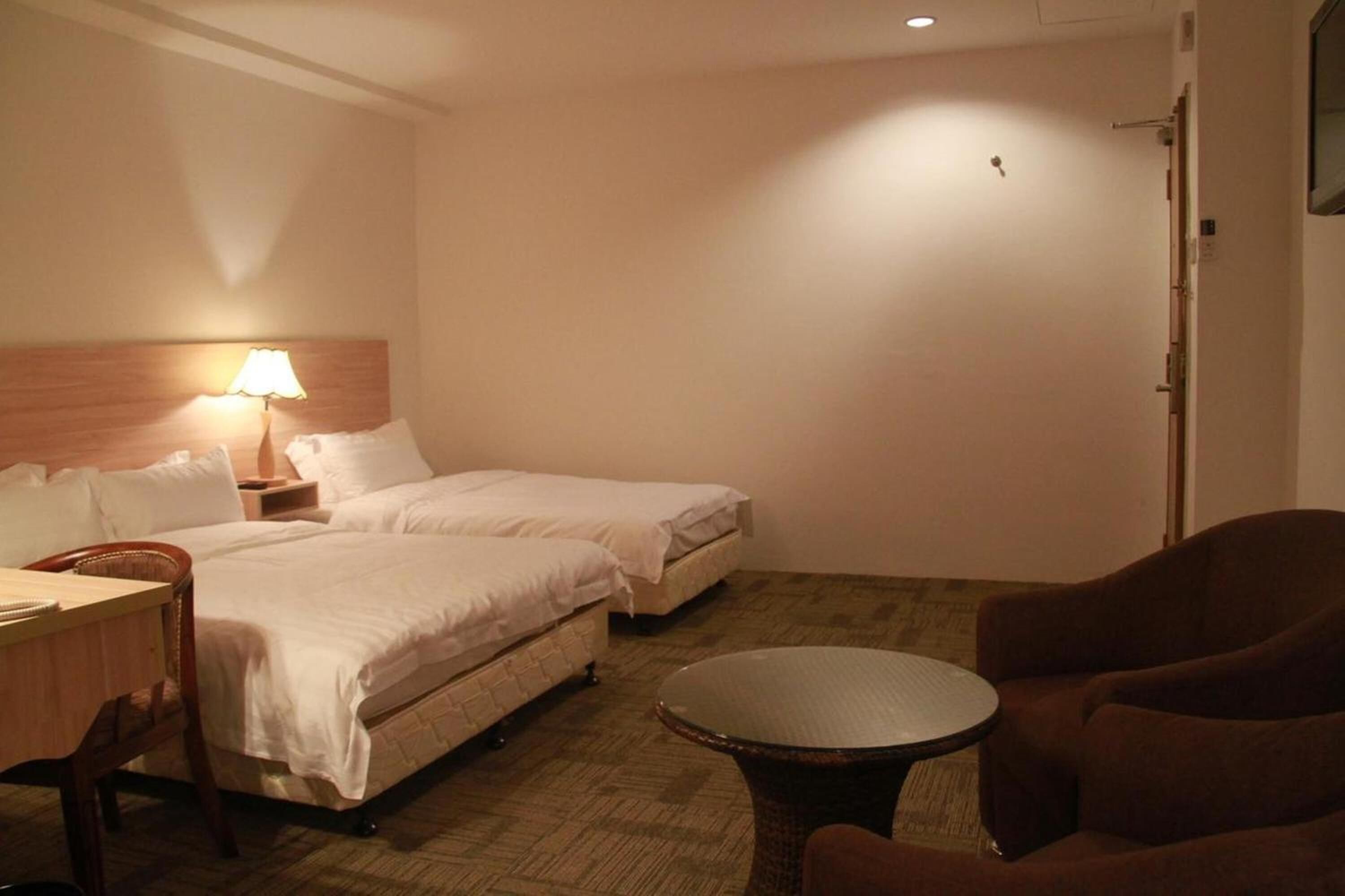 A hotel room with two beds, a small table, a chair, and a lamp.