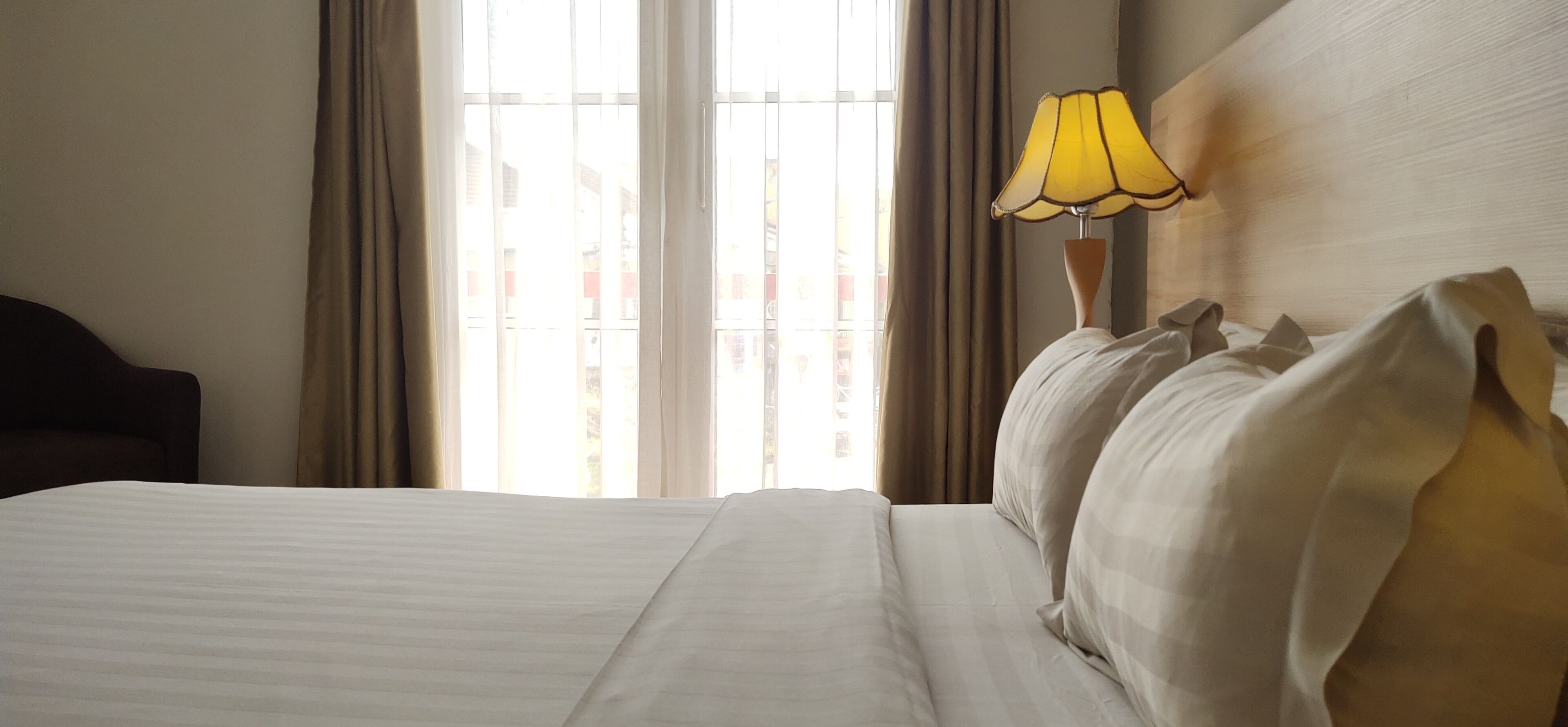 Habitación de hotel con cama, cortinas, lámpara y vistas a edificios a través del ventanal.