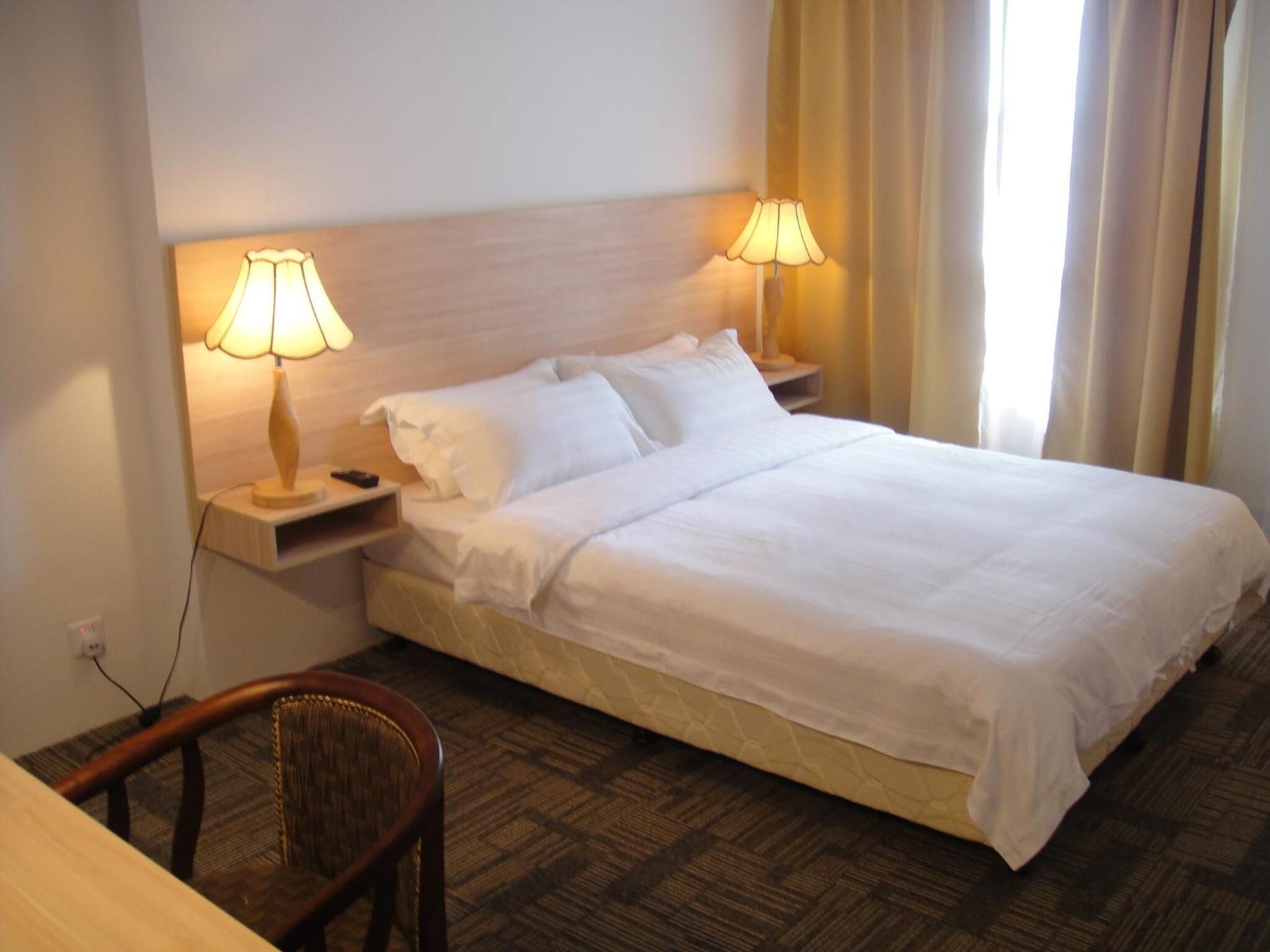 A hotel room with a bed, two bedside lamps, a nightstand, a chair, and a window with curtains.