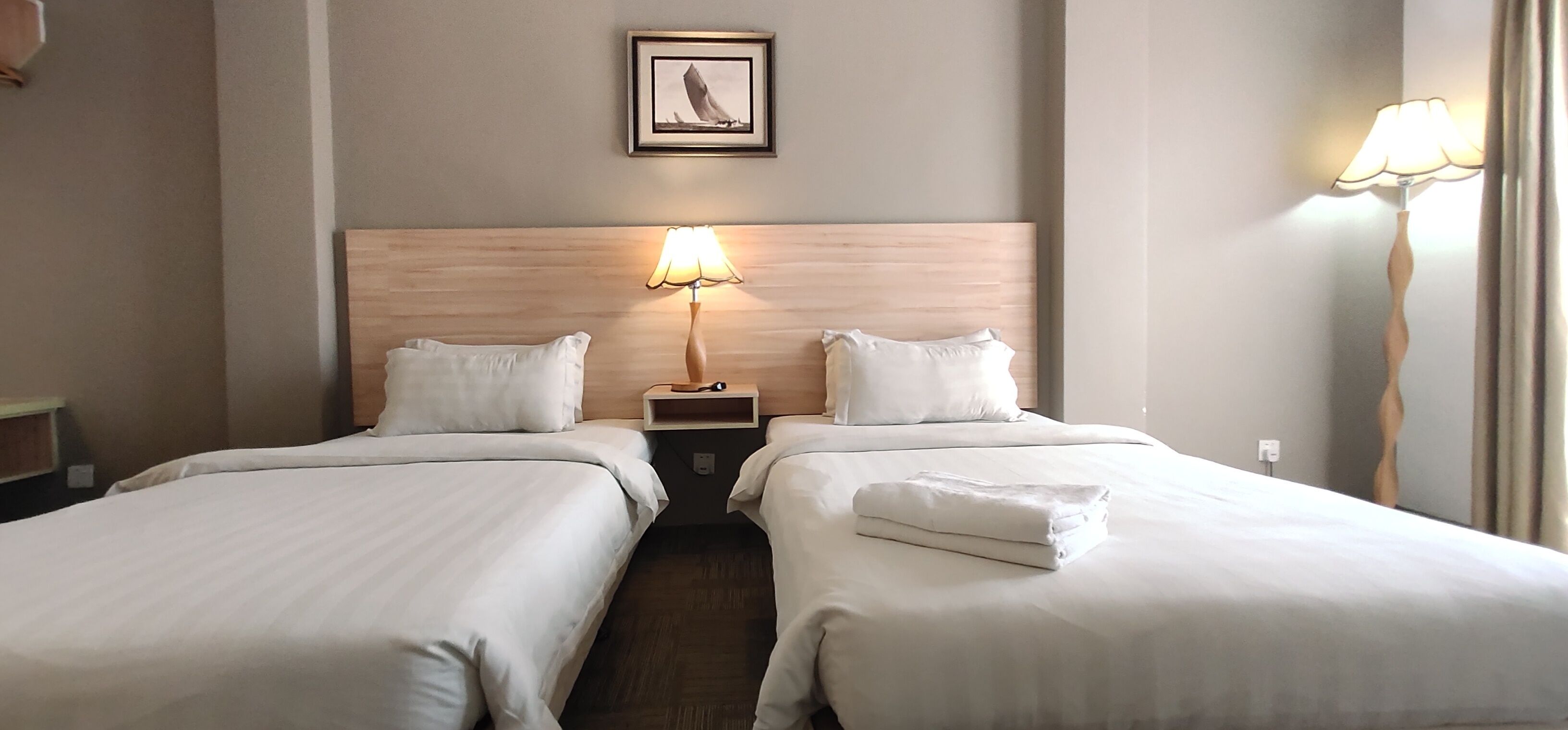 A hotel room with two beds, a wooden headboard, a bedside table with a lamp, and a framed picture on the wall.