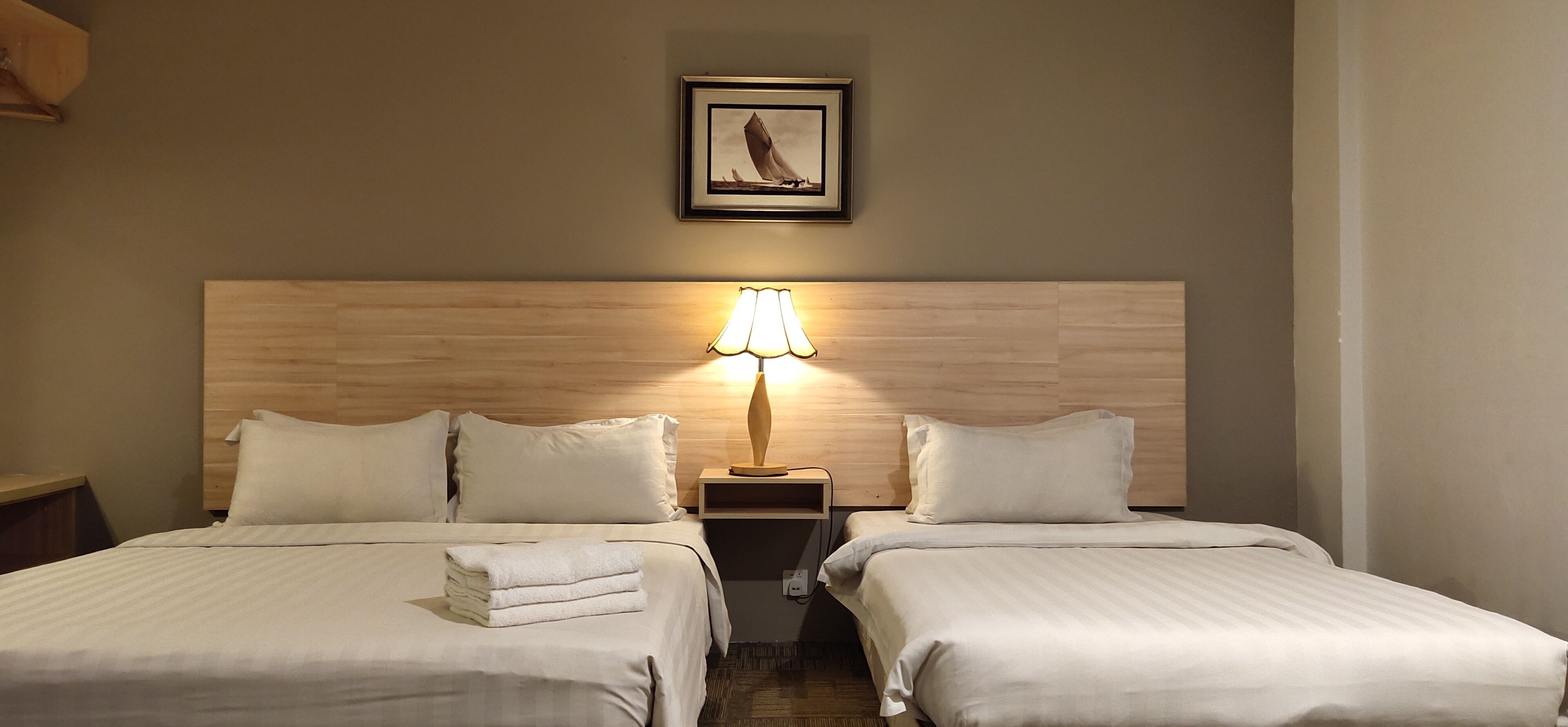 Habitación de hotel con dos camas, cabecera de madera, mesita de noche con lámpara y un cuadro en la pared.