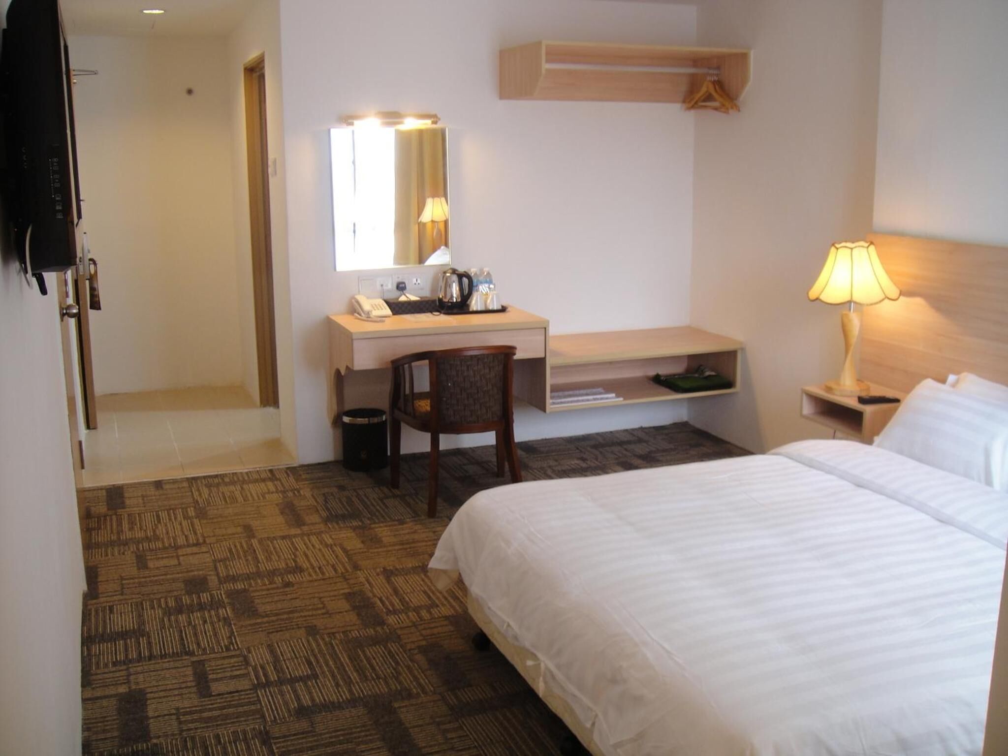 Une chambre d’hôtel comprenant un lit, un bureau avec une chaise, une table de chevet avec une lampe, un miroir et une étagère au-dessus du bureau.