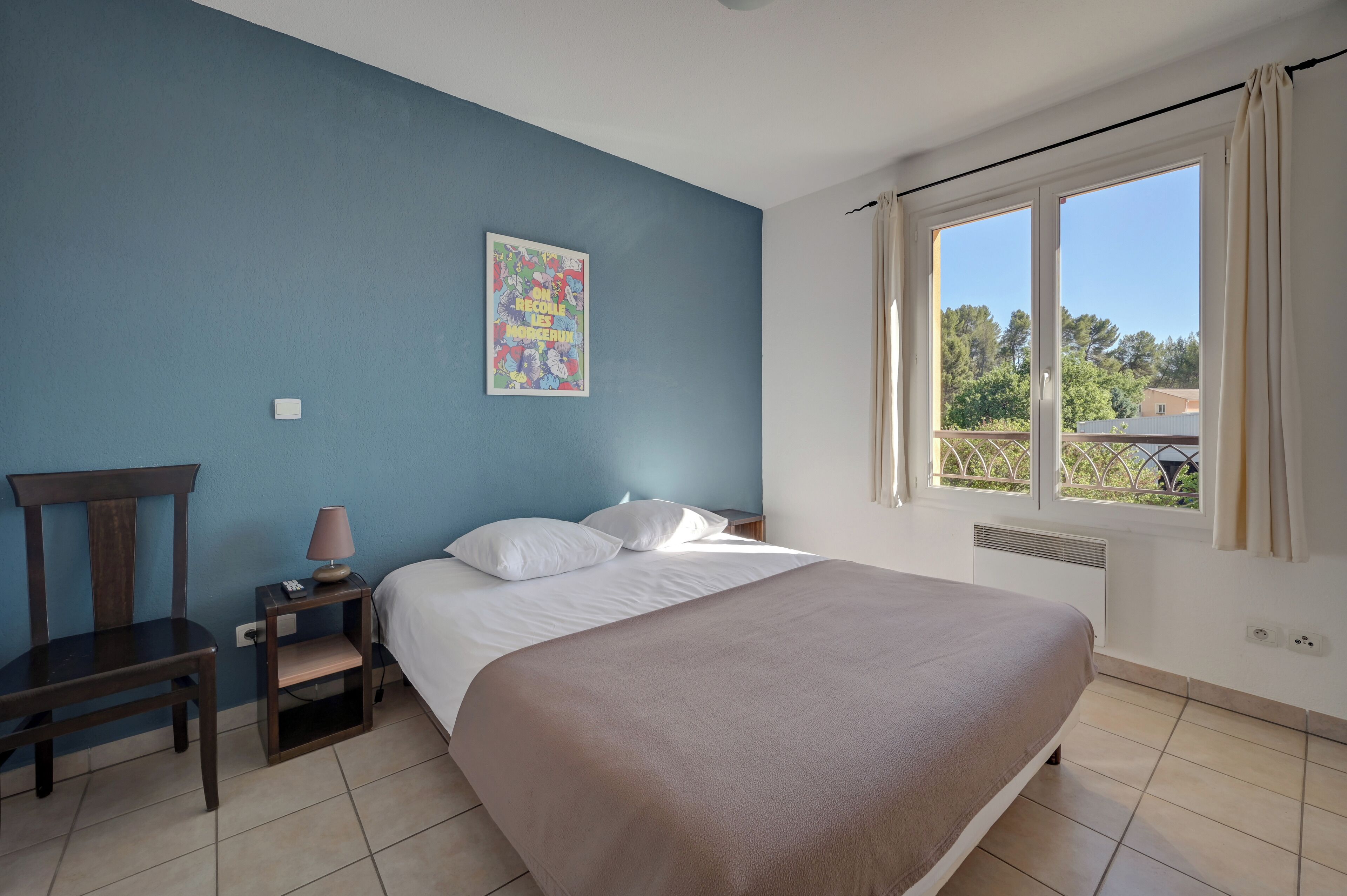 A bedroom with a bed, a chair, a nightstand, a lamp, a window with curtains, and a wall with a colorful poster.