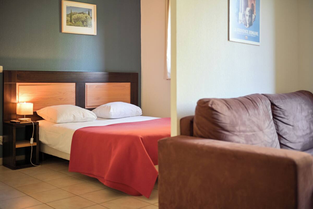 A hotel room with a bed, a nightstand with a lamp, a brown armchair, and a poster on the wall.