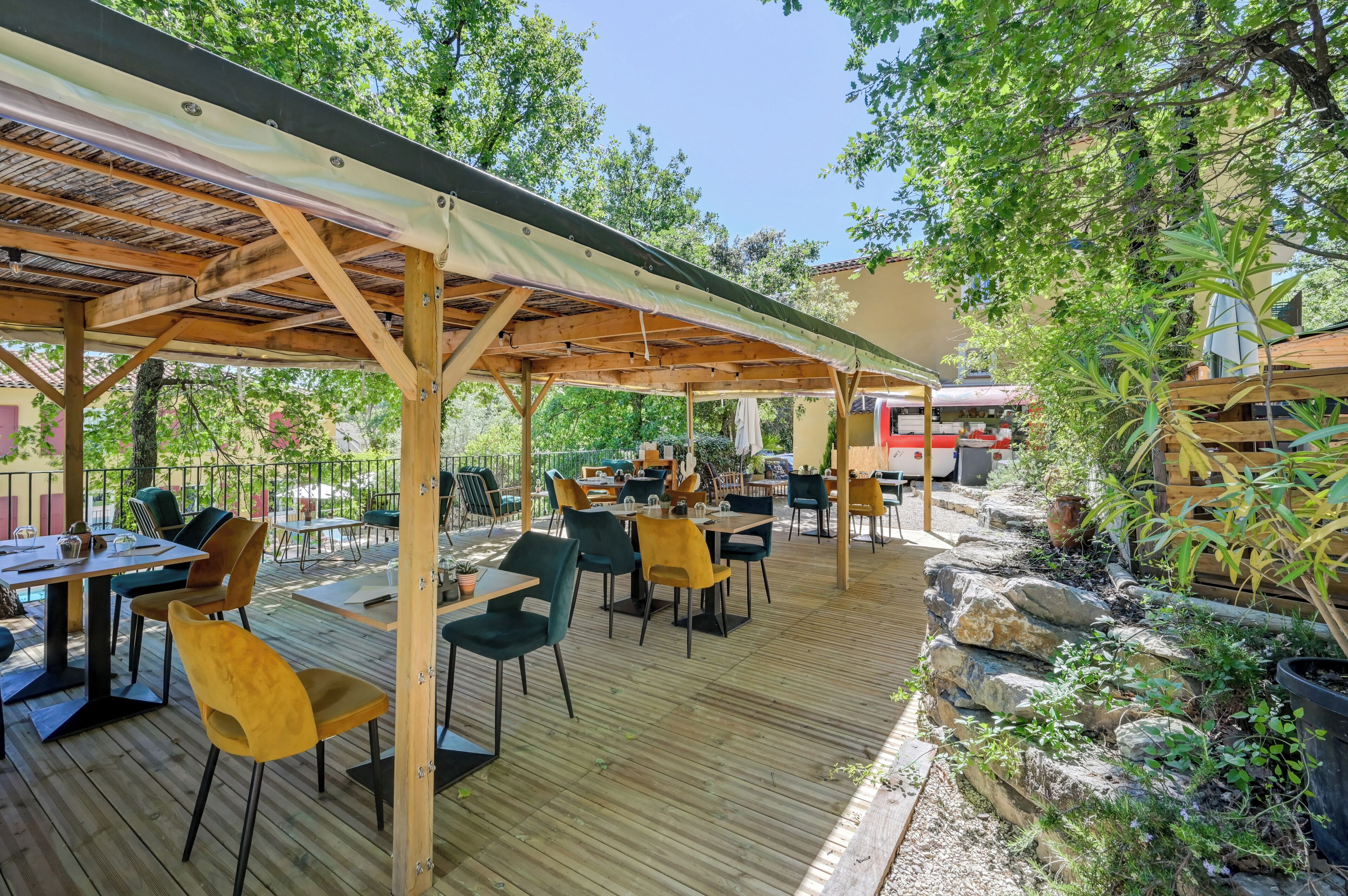 Une terrasse extérieure avec des tables et des chaises en bois, entourée de verdure et dotée d’un espace abrité.