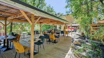 Une terrasse extérieure avec des tables et des chaises en bois, entourée de verdure et dotée d’un espace abrité.