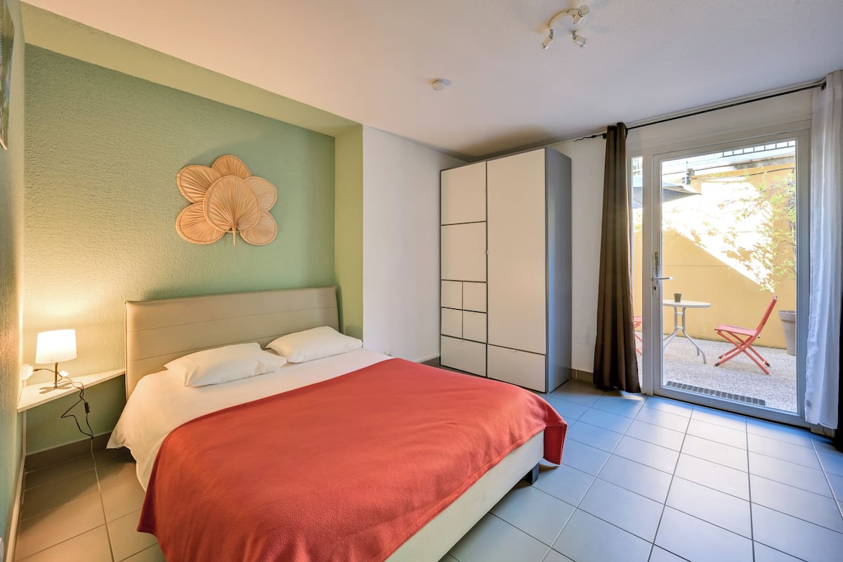 A hotel room with a bed, a nightstand, a wardrobe, and a sliding glass door leading to an outdoor seating area.