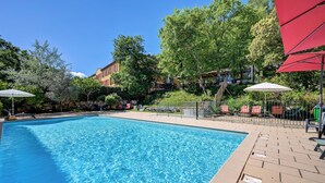 A clear blue pool surrounded by lounge chairs and umbrellas, with trees and a building in the background.