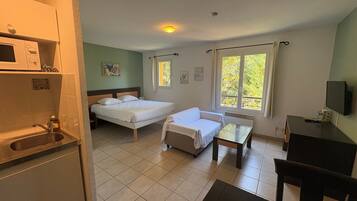A hotel room with a bed, a bedside table, a chair, a small desk, a TV, a window with curtains, a microwave, and a sink.