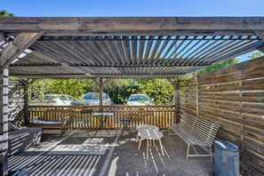 Een terras buiten met een houten pergola, zitgedeeltes en uitzicht op geparkeerde auto's.