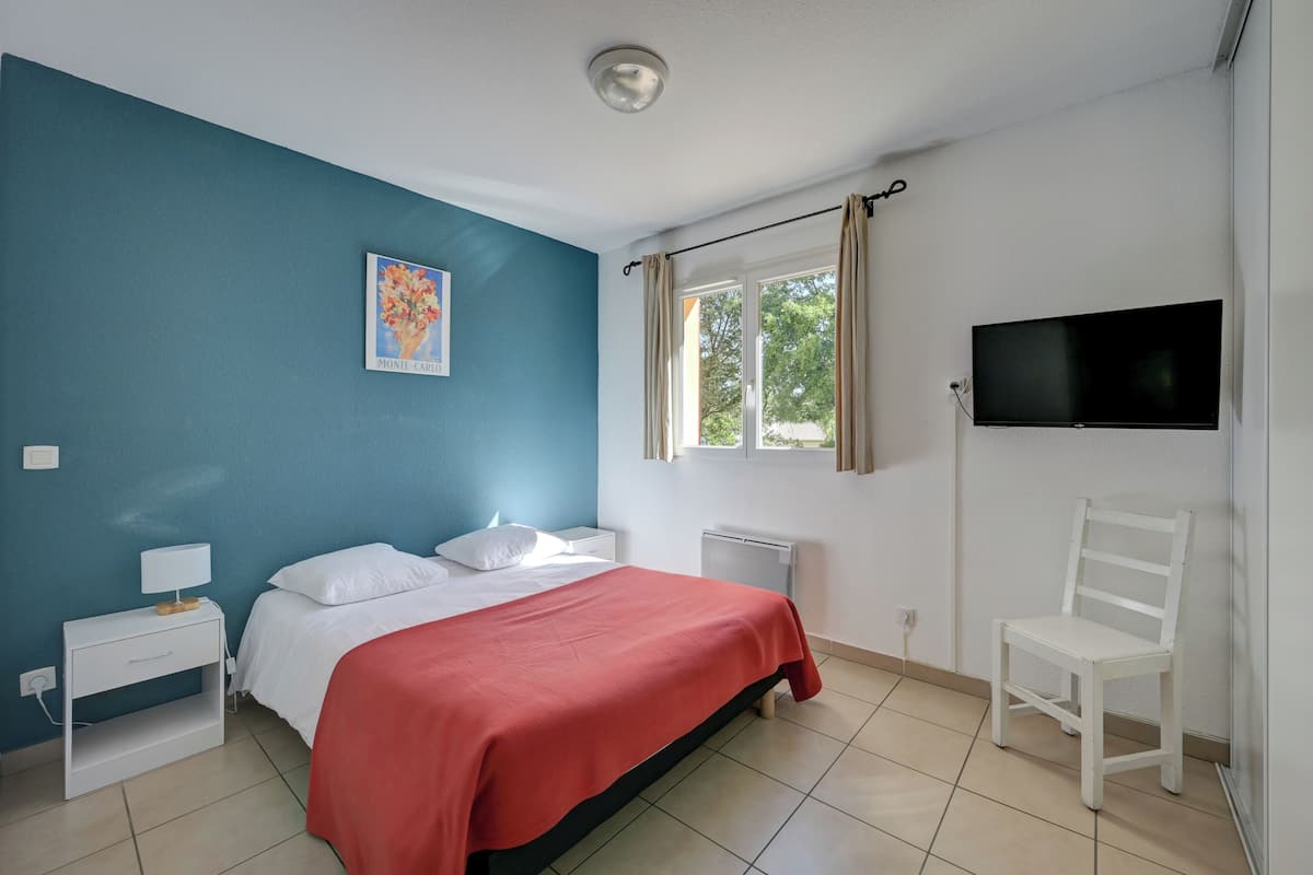 A bedroom with a bed, a nightstand, a chair, a TV, and a window with curtains.