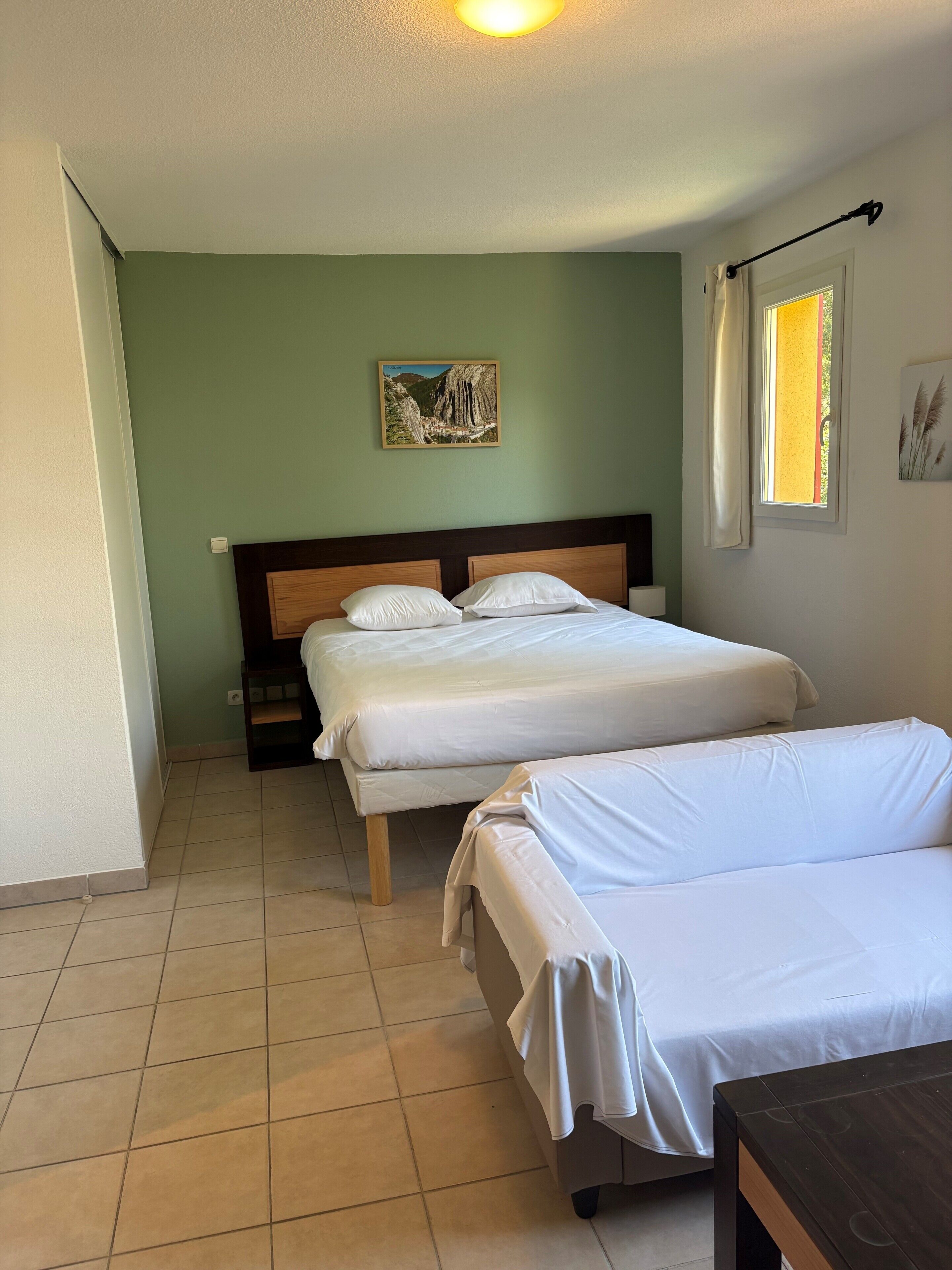 Una habitació amb dos llits, un panell de capçalera de fusta, un quadre a la paret, una finestra amb cortines i un sòl de rajola.